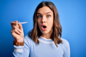 Young blonde girl holding dental orthodontic clear aligner over blue isolated background scared in shock with a surprise face, afraid and excited with fear expression